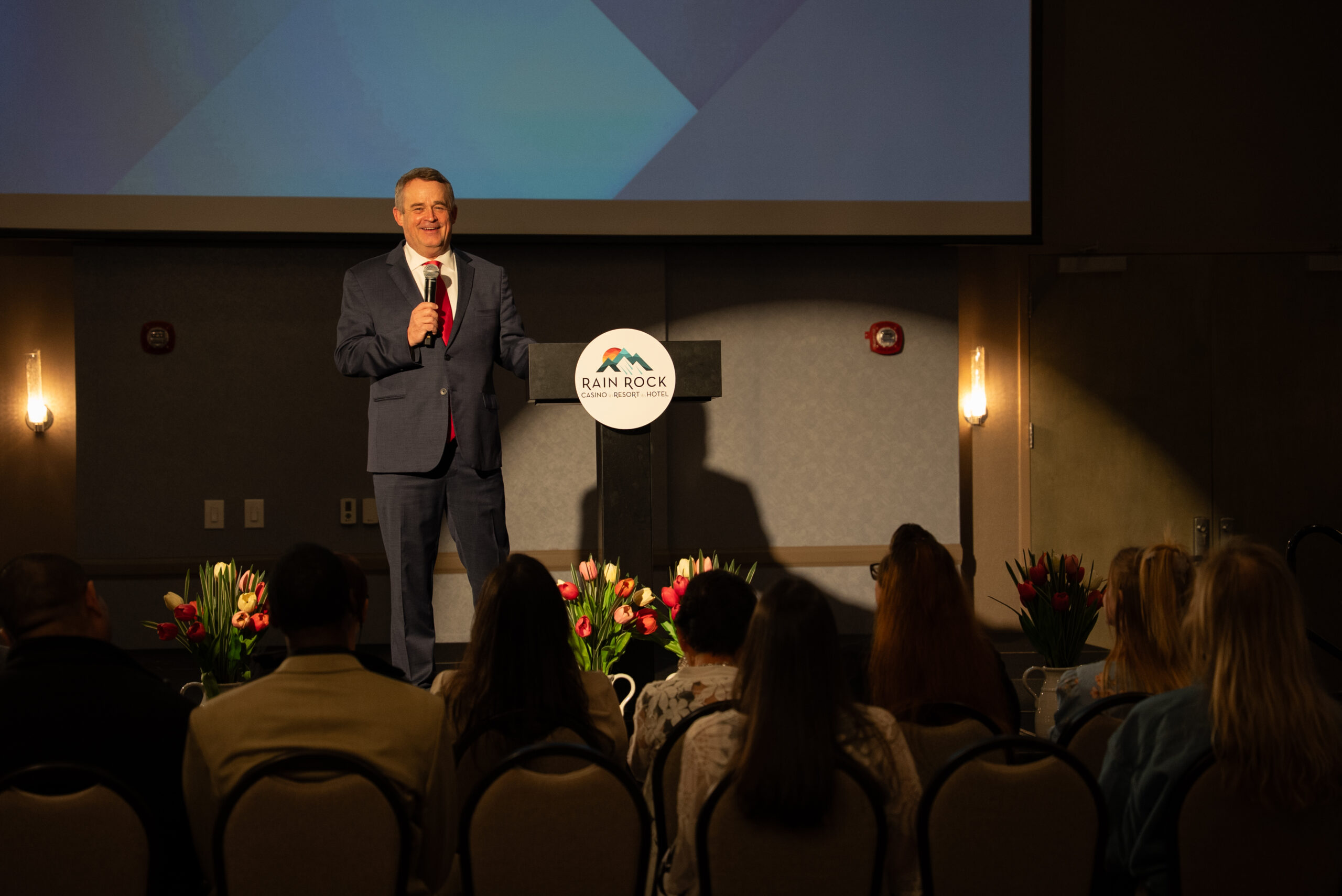 Speaker presenting on stage at a conference podium before a seated audience
